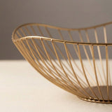 A close-up of a gold wire bowl with a curved, organic shape, resting on a light beige surface against a neutral gray background.