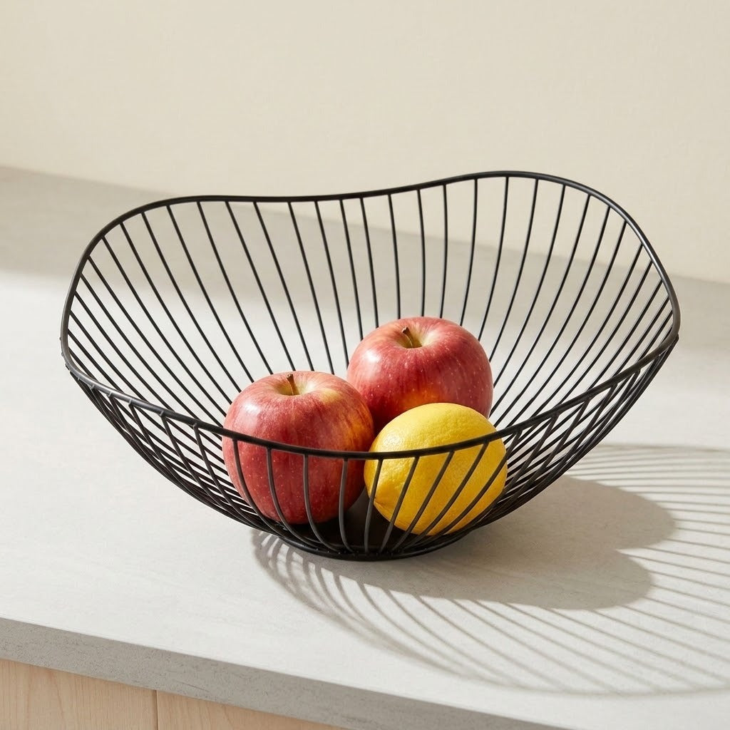 A black wire fruit basket with an organic, wavy shape sits on a light gray countertop, holding two red apples and a yellow lemon. The basket is photographed in a clean kitchen-like setting with soft natural light casting a shadow on the counter.