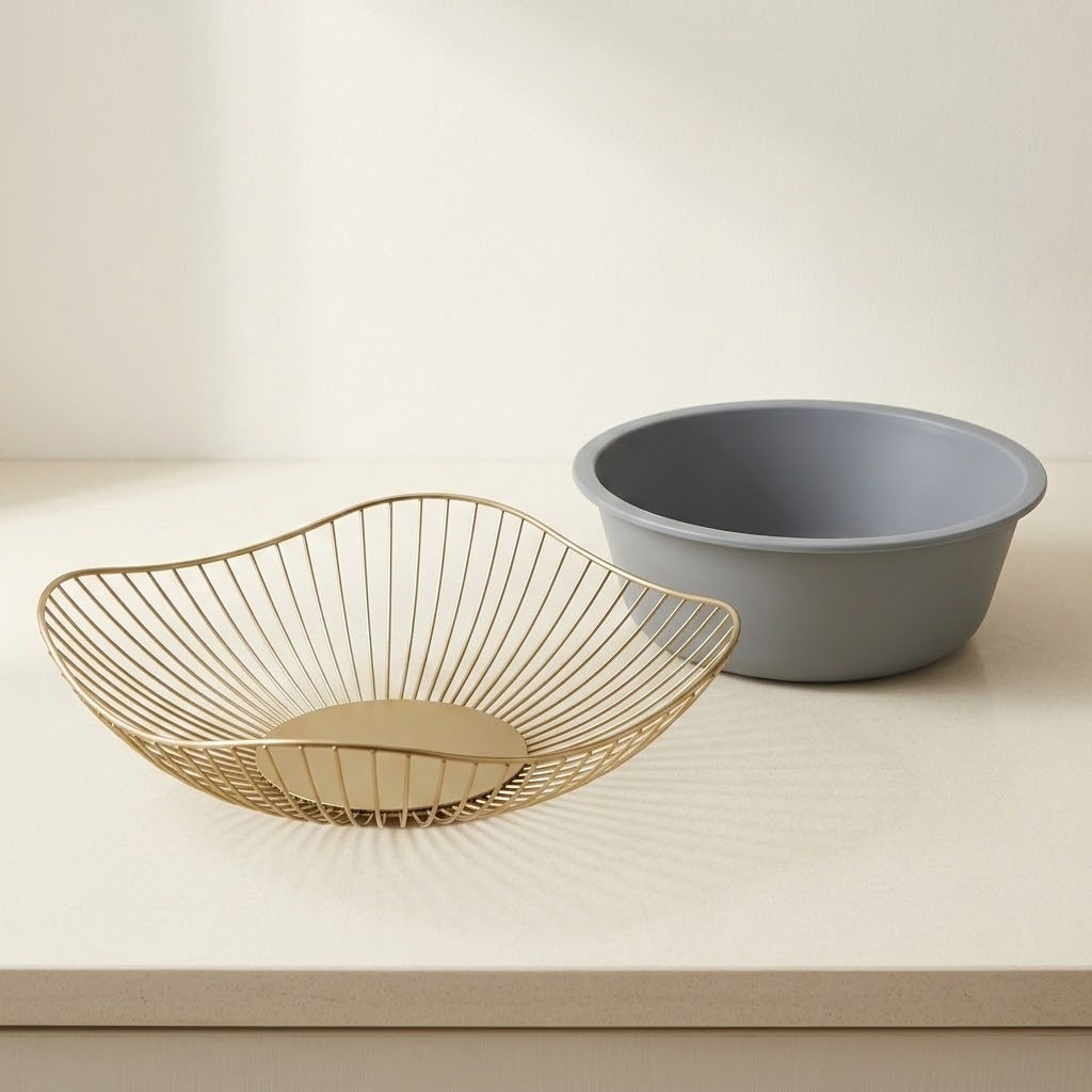 A gold wire bowl with a circular base sits in front of a matte gray plastic bowl on a light beige countertop against a cream background.