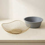 A gold wire bowl with a circular base sits in front of a matte gray plastic bowl on a light beige countertop against a cream background.