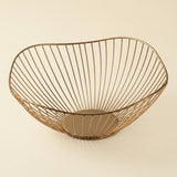 A modern wire bowl with a slatted design in a warm metallic gold finish, set against a solid cream background.
