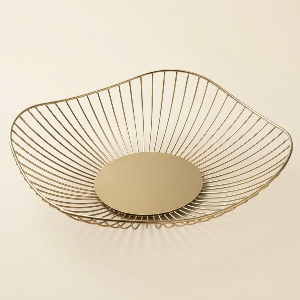 A modern wire bowl basket in a brushed gold finish, featuring a circular base and a slatted open top, shown on a solid light beige background.