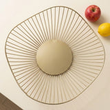 A modern oval wire bowl with a gold-toned metal frame and a round beige insert, photographed from above on a light cream surface with an apple and lemon in the corner.