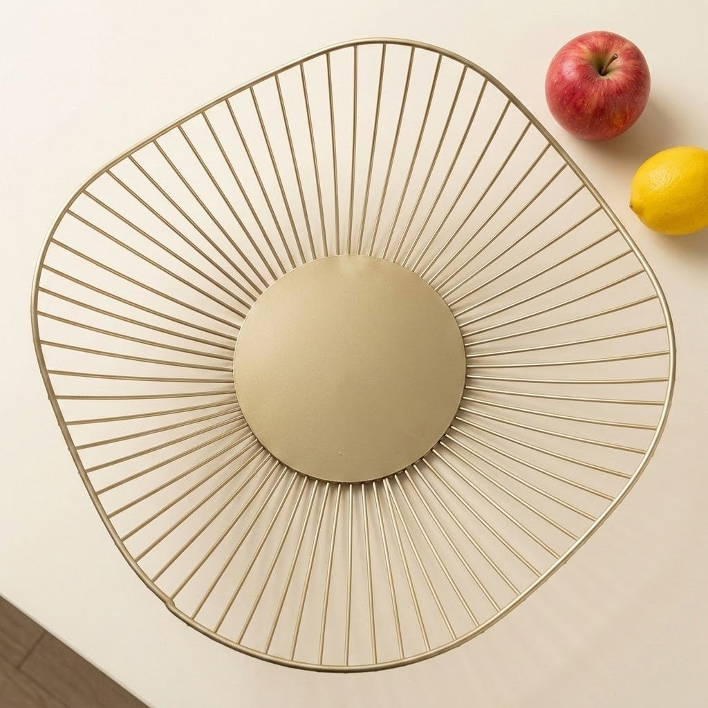 A modern oval wire bowl with a gold-toned metal frame and a round beige insert, photographed from above on a light cream surface with an apple and lemon in the corner.
