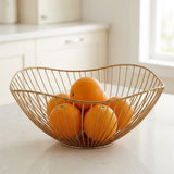 A gold wire fruit bowl filled with oranges on a white kitchen countertop, with a softly blurred kitchen background.