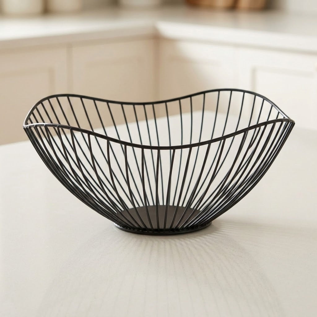 A black wire bowl with a curved, organic shape, displayed on a light-colored countertop with a softly blurred kitchen background.