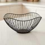 A black wire bowl with a curved, organic shape, displayed on a light-colored countertop with a softly blurred kitchen background.