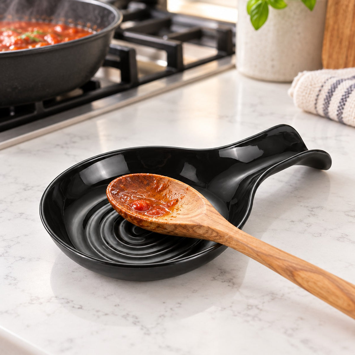 Black plastic spoon rest catches a sauce-coated spoon on a kitchen counter beside the stove.