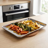 A rectangular stainless steel baking tray filled with roasted vegetables, including carrots, broccoli, and potato wedges, resting on a cork coaster on a light wood kitchen table with an oven in the background.