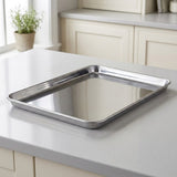 A rectangular stainless steel baking pan with raised edges sits on a light gray kitchen countertop, with cream cabinets and a small potted plant blurred in the background.
