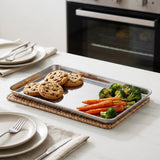 A rectangular stainless steel baking sheet resting on a woven placemat on a white table, holding chocolate chip cookies and roasted broccoli and carrots, with plates and an oven blurred in the background.