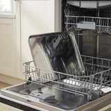 An open stainless steel dishwasher with the upper rack extended, holding a large reflective metal baking sheet and a gray dish rack insert, set in a kitchen environment.