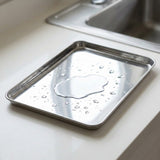 A square stainless steel tray with raised edges sits on a white kitchen countertop near a sink, with water droplets on the reflective surface.