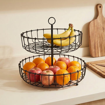 A two-tier black wire fruit basket on a white countertop, filled with oranges, apples, and bananas, with a wooden cutting board in the background.