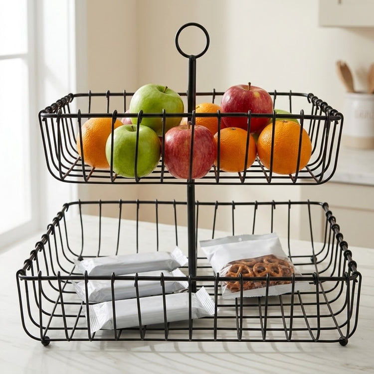 A two-tier black wire basket food caddy holding oranges, apples, and packaged snacks, set on a light countertop in a bright kitchen-like interior.
