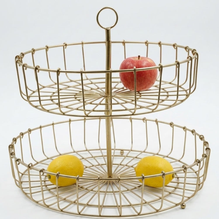 A two-tier round wire fruit stand in a brushed gold finish, shown with a red apple on the top tier and two yellow lemons on the bottom tier against a white background.