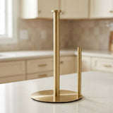 A modern two-stick paper towel holder with a round base in brushed gold, shown on a white marble kitchen countertop with a blurred kitchen background.