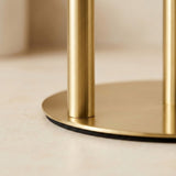 Close-up of a round gold metal base with two vertical gold cylindrical legs on a light beige surface.