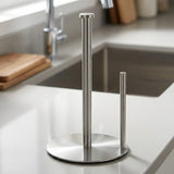 A brushed stainless steel paper towel holder with a tall main pole, a shorter side holder, and a round base, shown on a kitchen countertop near a sink.