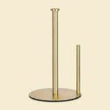 A modern brass paper towel or napkin holder with a round base, featuring one tall stand and one short stand, shown against a solid cream background.