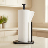 A black paper towel holder with a roll of white paper towels, set on a light beige kitchen countertop with a blurred kitchen background.