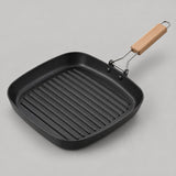 Square grill pan with ridged black cooking surface and light wood grip on a pale studio backdrop.