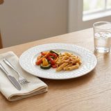A white textured ceramic plate with penne pasta and vegetables, set on a light wood table next to a folded cream napkin with a knife and fork, and a glass of water.