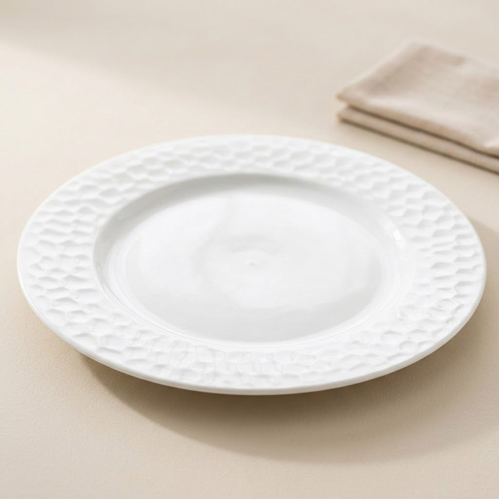 A white ceramic dinner plate with a textured, dimpled pattern around the rim, shown on a neutral beige surface with a folded napkin in the background.