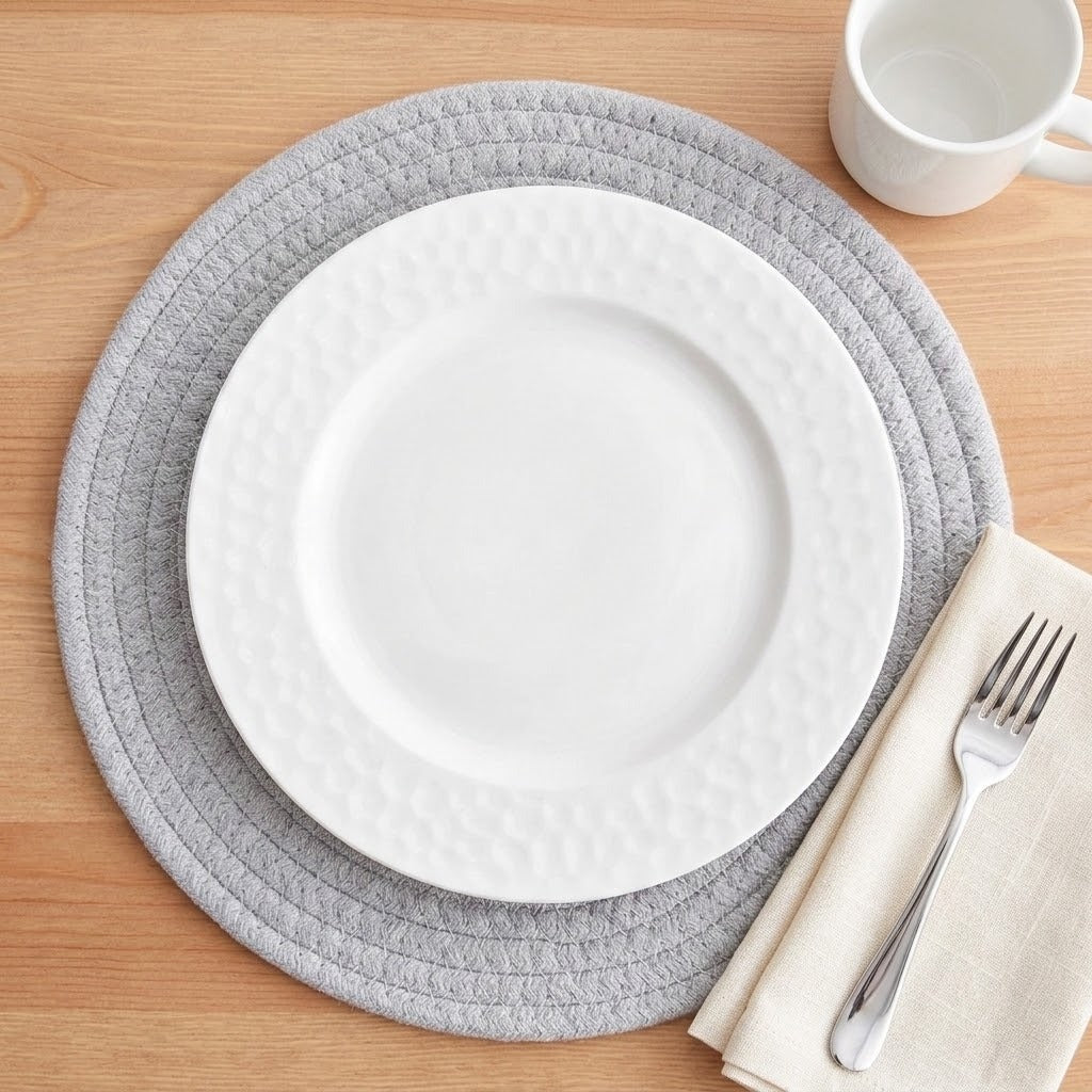 A place setting on a light wood table featuring a round gray braided placemat, a white textured ceramic plate, a folded cream napkin, a silver fork, and a white mug in the corner.