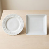 A white ceramic plate with a textured, dimpled border and a square white plate, displayed side-by-side on a light wood tabletop against a neutral beige background.