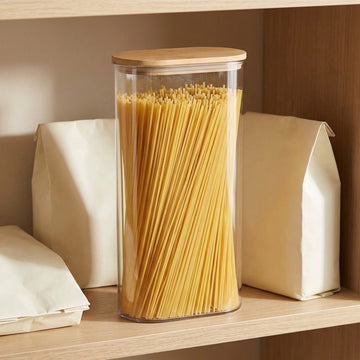 A tall clear cylindrical canister with a light wood lid, filled with spaghetti, sitting on a wooden shelf between cream paper bags.
