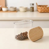 A clear plastic coffee canister filled with coffee beans, shown next to its light bamboo lid on a light beige countertop, with a softly blurred kitchen background.