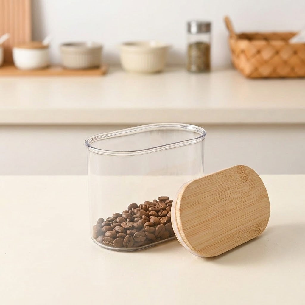 A clear plastic coffee canister filled with coffee beans, shown next to its light bamboo lid on a light beige countertop, with a softly blurred kitchen background.