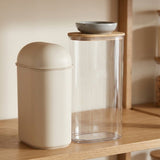 A beige capsule-shaped canister with a matching beige lid sits next to a tall clear glass jar with a light wood lid and a small gray bowl on top, displayed on a wooden shelf.