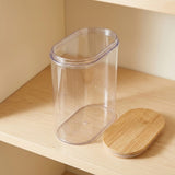 A clear plastic storage jar with an oval shape and a light natural wood lid, shown on a light wood shelf.