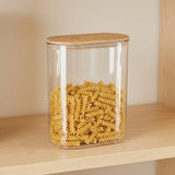 A clear cylindrical storage jar filled with rotini pasta, featuring a light natural wood lid, displayed on a wooden shelf against a beige wall.