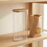 A tall clear cylindrical canister with a light wood lid sits on a wooden shelf next to a brown paper bag filled with penne pasta.
