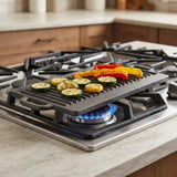 A black cast iron gas grill plate with ridges sits on a lit blue gas burner of a stainless steel gas stove, cooking sliced zucchini and bell peppers on a light beige marble kitchen countertop.