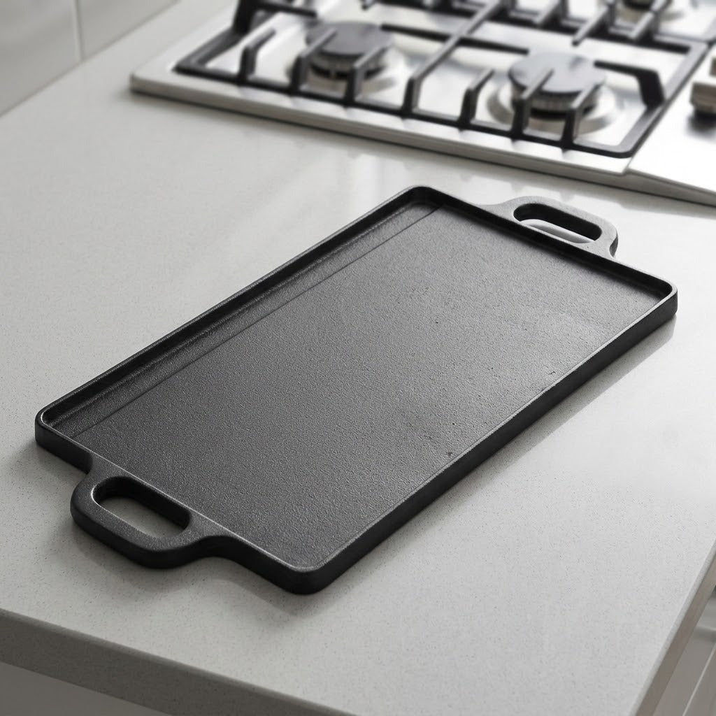A rectangular black cast iron griddle or flat-top pan with integrated side handles, resting on a light gray kitchen countertop with a gas stovetop visible in the background.