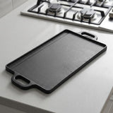 A rectangular black cast iron griddle or flat-top pan with integrated side handles, resting on a light gray kitchen countertop with a gas stovetop visible in the background.