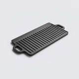 A rectangular black cast iron griddle plate with two loop handles, shown at an angle on a light gray background.