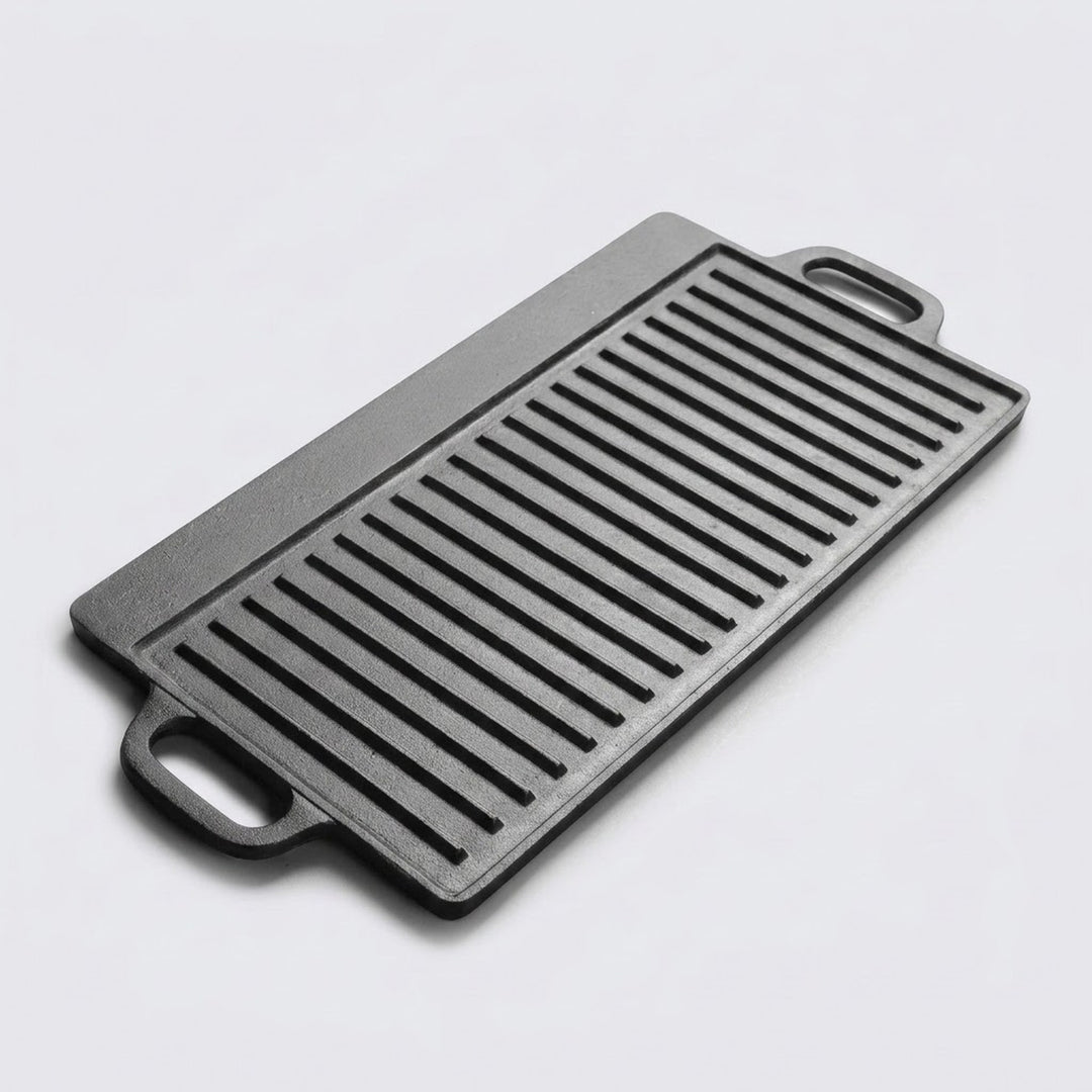 A rectangular black cast iron griddle plate with two handles and parallel ridges, photographed on a light gray background.