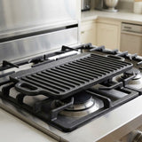 A black cast iron griddle with ridges and built-in handles, shown resting on a stainless steel gas stovetop in a kitchen setting.