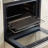 A dark cast iron grill pan with side handles is shown on a metal oven rack inside an open built-in oven. Cream-colored kitchen cabinets are visible on both sides.
