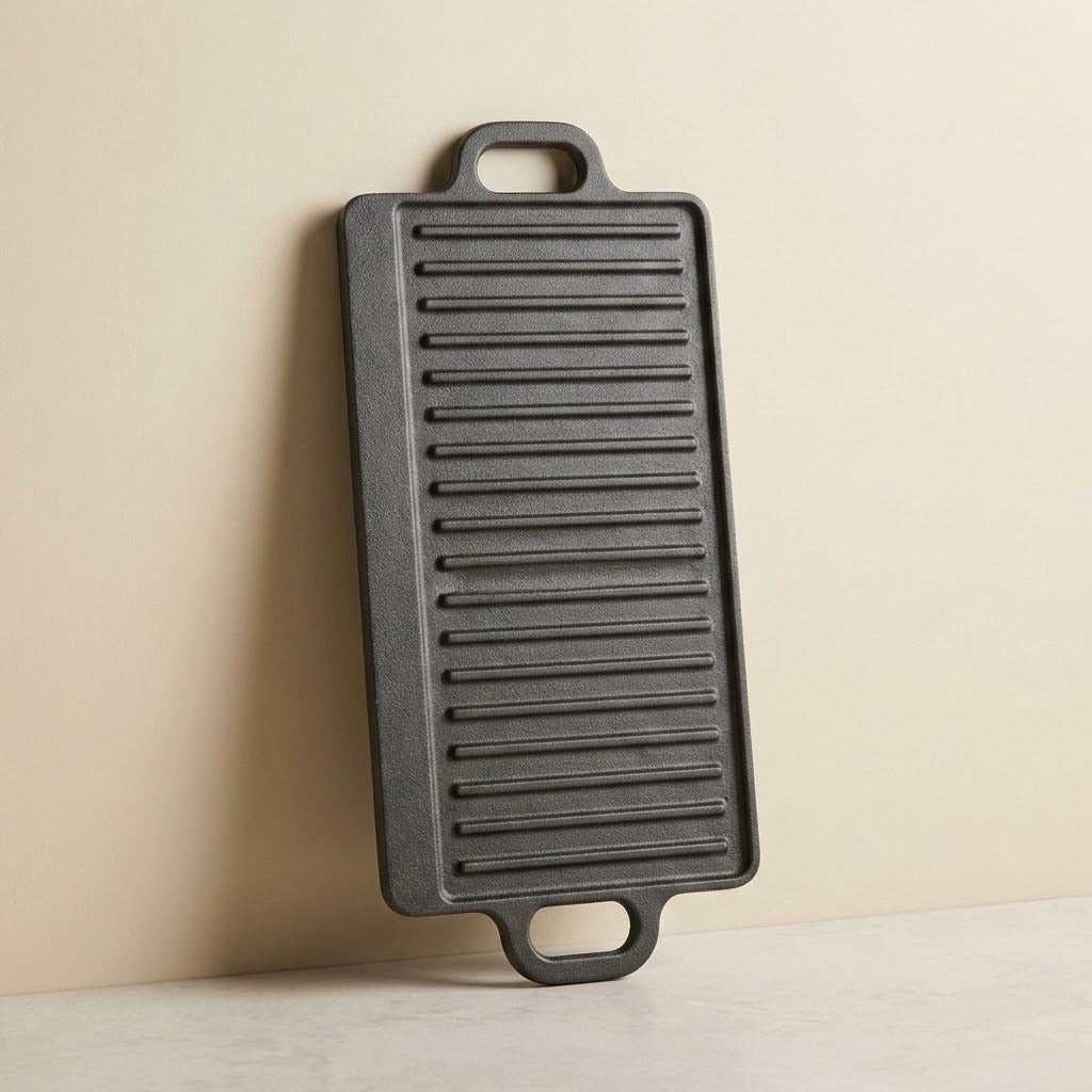 A dark gray cast iron rectangular griddle with ridges and two handles, leaning against a beige wall on a light surface.