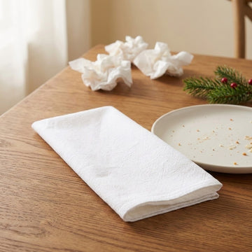A folded white cotton napkin on a wooden table next to a plate, with crumpled napkins and a green pine sprig in the background.