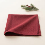 A folded deep burgundy red linen napkin on a light cream background, styled with a small green pine branch and red berries in the top right corner.