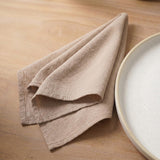 A folded beige linen cloth napkin or tea towel on a light wood tabletop next to a white speckled ceramic plate.