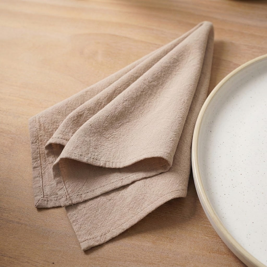 A folded beige linen cloth napkin or tea towel on a light wood tabletop next to a white speckled ceramic plate.
