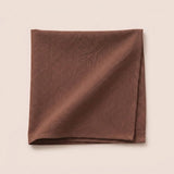 A folded brown fabric cloth, likely a napkin or pocket square, displayed flat against a solid, warm off-white background.